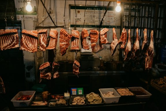 Fleischstücken hängen an einem Markstand, daneben verschiedene Fleischwaren in Behältern.